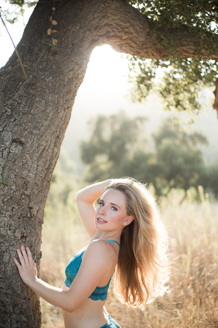 Woman with long hair in a blue lace top poses beside a tree in a sunlit field.