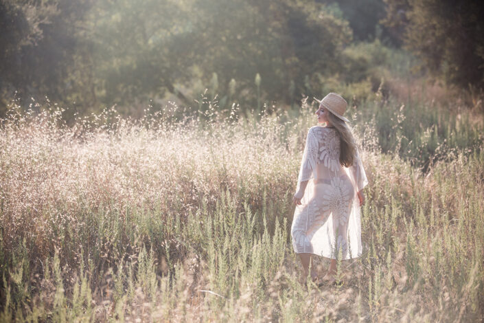 A woman in a light, lacy outfit and a straw hat walks through a sunlit field of tall grass.
